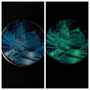Peut inclure: Une assiette décorative ronde représentant un grand voilier naviguant sur une mer orageuse sous une grande lune. L'œuvre est principalement bleue avec une bordure dorée. L'image brille dans le noir.