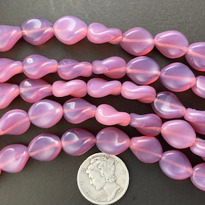 May include: A strand of pink glass beads, each bead is a unique, irregular shape. The beads are translucent and have a slight shimmer. The beads are arranged in a loose pile, with a few beads overlapping. A silver coin is in the center of the image for scale.
