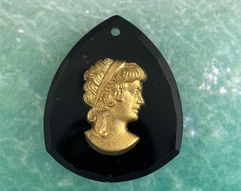 May include: A black cameo pendant featuring a gold-coloured profile of a woman wearing a headband. The pendant is teardrop-shaped and has a hole at the top for hanging.