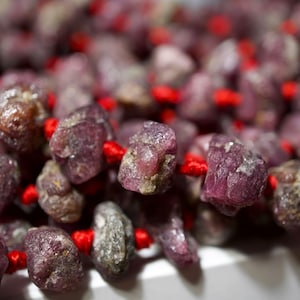 May include: A close-up of a strand of pink gemstone beads strung on red thread. The beads are irregular in shape and have a slightly rough texture.