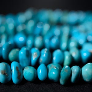 May include: Close-up of turquoise gemstone beads. The beads are a vibrant turquoise color with variations in shade and texture. The beads are strung together, creating a necklace or bracelet. The background is dark, which makes the turquoise color pop.