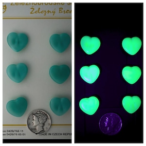 May include: Six teal heart-shaped buttons arranged in two columns on a white card, shown in daylight and glowing green in the dark. A silver Mercury dime is placed next to the bottom left button for scale, with text visible at the top and bottom.