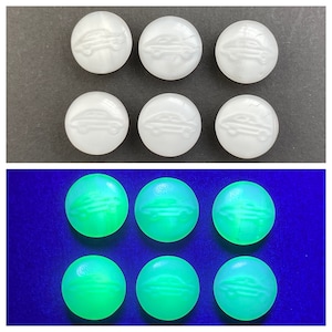 May include: Six white buttons with a car design, shown under a black light, glowing green.