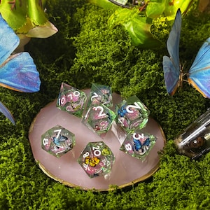 Può includere: Un set di sette dadi poliedrici in resina trasparente con ali di farfalla in miniatura e glitter rosa incorporati, caratterizzati da numeri bianchi. I dadi sono disposti su un sottobicchiere in pietra rosa con un bordo metallico dorato.