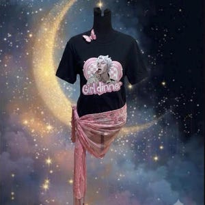 May include: A black short-sleeve t-shirt featuring a graphic of a person with white hair eating, surrounded by a pink heart and checkered pattern, with the text Girl dinner. A small pink butterfly is attached to the neckline, and a pink scarf is tied.