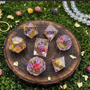 May include: A set of seven polyhedral dice made of clear resin, each containing small dried flowers in shades of pink and yellow, along with gold leaf flakes. The numbers on the dice are painted in a light purple color, featuring various geometric shapes.