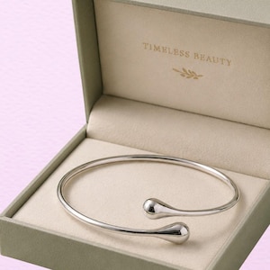 May include: A silver-toned open cuff bracelet with rounded, teardrop-shaped ends resting inside a sage green hinged box. The interior of the box lid features the gold-colored text "TIMELESS BEAUTY" accompanied by a small leaf icon.
