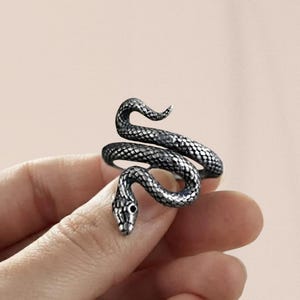 May include: A silver-toned metal ring shaped like a coiled snake. The ring features a detailed, textured scale pattern across its entire surface, with a defined head and a curved tail that wraps around to form the band of the jewelry piece.