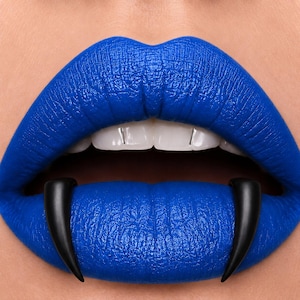 May include: A pair of smooth, curved black fangs positioned over the lower lip, which is coated in a vibrant, matte royal blue lipstick. The upper lip is also painted in the same intense blue shade, revealing white teeth with small silver accents.