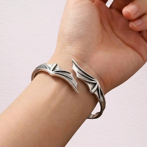 May include: A silver-toned cuff bracelet featuring a design of two symmetrical bat wings that meet at the center. The metal has a polished finish with recessed details that highlight the structure of the wing membranes and the pointed tips.
