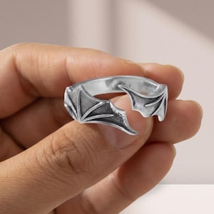 Könnte beinhalten: Ein silberfarbener offener Ring mit einem Design aus zwei symmetrischen Fledermausflügeln, die sich in der Mitte treffen. Das Metall hat eine polierte Oberfläche mit abgedunkelten Vertiefungen, um die Textur der Flügel hervorzuheben.
