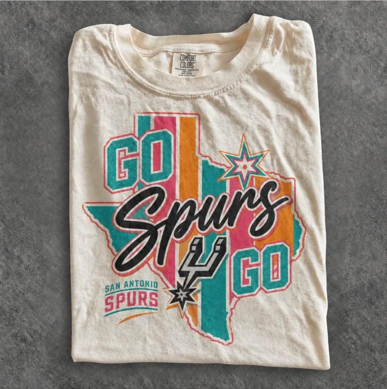 May include: A cream-colored t-shirt featuring a graphic of the state of Texas in teal, pink, and orange stripes. The text reads GO SPURS GO with the San Antonio Spurs logo and the words SAN ANTONIO SPURS printed in teal and pink lettering.