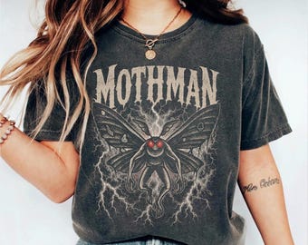 Puede incluir: Una camiseta gris carbón de manga corta con un estampado gráfico del Mothman con alas grandes y ojos rojos brillantes. El texto MOTHMAN está impreso en una fuente desgastada sobre la criatura, rodeado de ilustraciones de rayos.