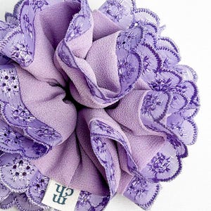 Puede incluir: Un coletero de color lavanda con una base de tela fruncida y un borde de encaje festoneado con intrincados bordados florales. Una pequeña etiqueta de tela blanca con las letras negras "ch" está cosida en el lateral del coletero.