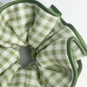 Puede incluir: Un coletero de tela con un patrón de cuadros vichy en verde claro y blanco. El accesorio está rematado con un borde ribeteado en verde oscuro sólido a lo largo del perímetro fruncido, creando una apariencia texturizada y recogida.