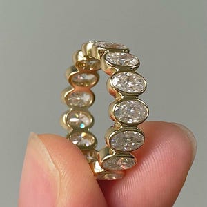 Puede incluir: Un anillo de oro con una serie de diamantes de talla ovalada engastados en una banda continua. Los diamantes son claros y brillantes, y el anillo tiene un acabado pulido en oro amarillo. El anillo está sostenido por los dedos de una persona.