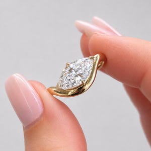 May include: A gold ring featuring a marquise-cut clear gemstone set in a polished yellow gold band. The stone is held by prongs and the band curves around the base of the setting, creating a sleek, continuous metal frame around the center stone.