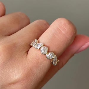 Puede incluir: Un anillo de oro con una serie de piedras preciosas claras de diferentes formas, incluyendo óvalos, esmeraldas y cortes de pera. El anillo está engastado en una banda de oro amarillo y se lleva en un dedo.