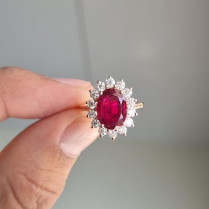 May include: A gold ring featuring a large, oval-cut, deep red ruby at the centre, surrounded by a halo of twelve small, round, clear white diamonds. The band is a polished gold metal, and the setting creates a floral-like cluster design.