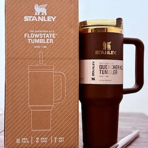 May include: A brown Stanley Flowstate tumbler with a handle, straw, and gold-colored lid, next to its cardboard box. The box and tumbler have the Stanley logo and text that reads "40 OZ | 1.18L". The tumbler keeps drinks cold for 2 days.