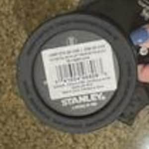 Op de afbeelding: Een donkergrijze Stanley merkdeksel met een witte sticker. De sticker heeft een barcode en tekst. De deksel is rond en gemaakt van plastic. De deksel ligt op een bruine ondergrond.