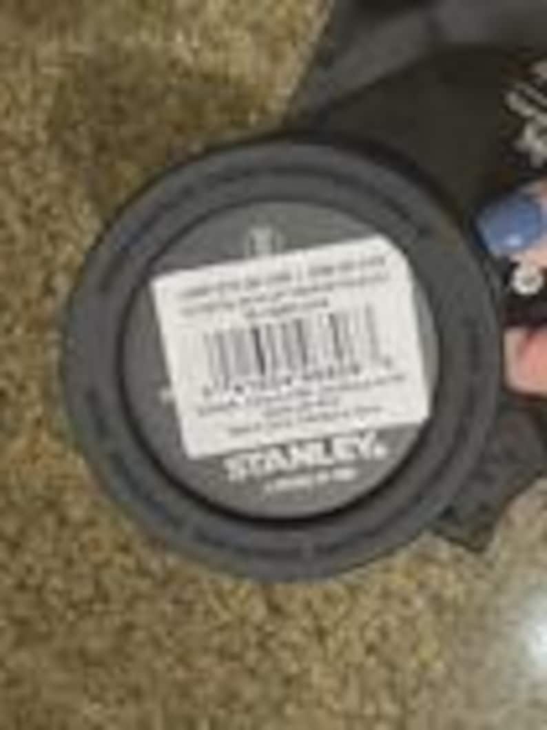 Op de afbeelding: Een donkergrijze Stanley merkdeksel met een witte sticker. De sticker heeft een barcode en tekst. De deksel is rond en gemaakt van plastic. De deksel ligt op een bruine ondergrond.
