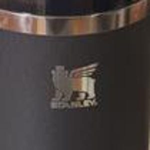 Op de afbeelding: Een zwarte Stanley thermosfles met een zilveren rand en het Stanley-logo. Het logo toont een gevleugelde leeuw en de merknaam. De thermosfles is ontworpen om dranken warm of koud te houden.