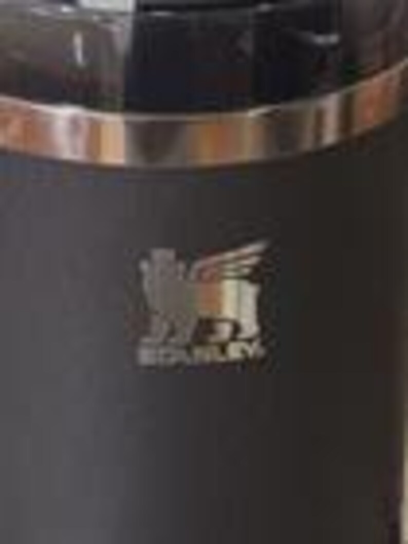 Op de afbeelding: Een zwarte Stanley thermosfles met een zilveren rand en het Stanley-logo. Het logo toont een gevleugelde leeuw en de merknaam. De thermosfles is ontworpen om dranken warm of koud te houden.