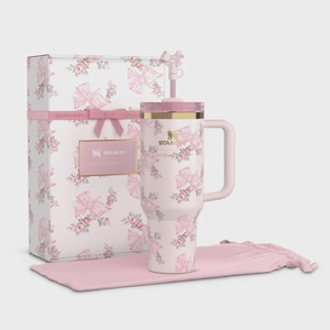 Puede incluir: Un vaso Stanley Lovesha blanco con un estampado de lazo y flores rosas, una caja de regalo a juego y una funda de transporte rosa. El vaso tiene un asa y una pajita. La caja de regalo est&aacute; decorada con lazos y rosas rosas. El logotipo de Stanley es visible.