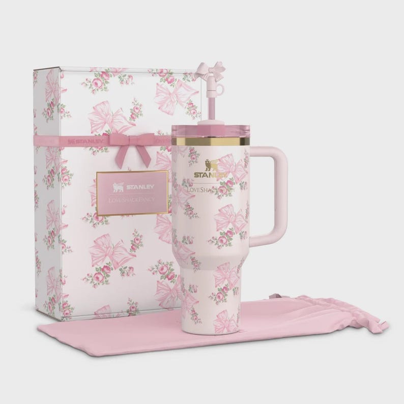 Puede incluir: Un vaso Stanley Lovesha blanco con un estampado de lazo y flores rosas, una caja de regalo a juego y una funda de transporte rosa. El vaso tiene un asa y una pajita. La caja de regalo est&aacute; decorada con lazos y rosas rosas. El logotipo de Stanley es visible.