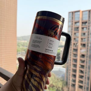 May include: A stainless steel tumbler with a black handle and lid. The tumbler features a pattern of gold and brown stripes and a red and brown design. The tumbler has a label that reads "FLOWSTATE - POSITION LID".