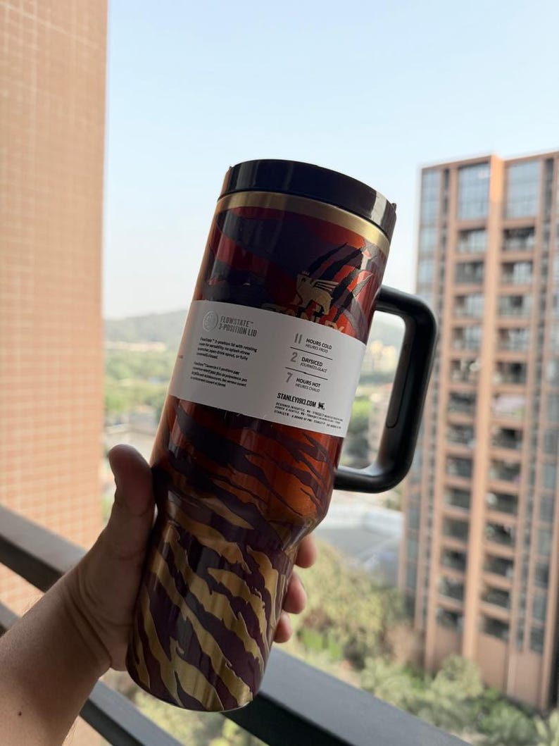 May include: A stainless steel tumbler with a black handle and lid. The tumbler features a pattern of gold and brown stripes and a red and brown design. The tumbler has a label that reads "FLOWSTATE - POSITION LID".