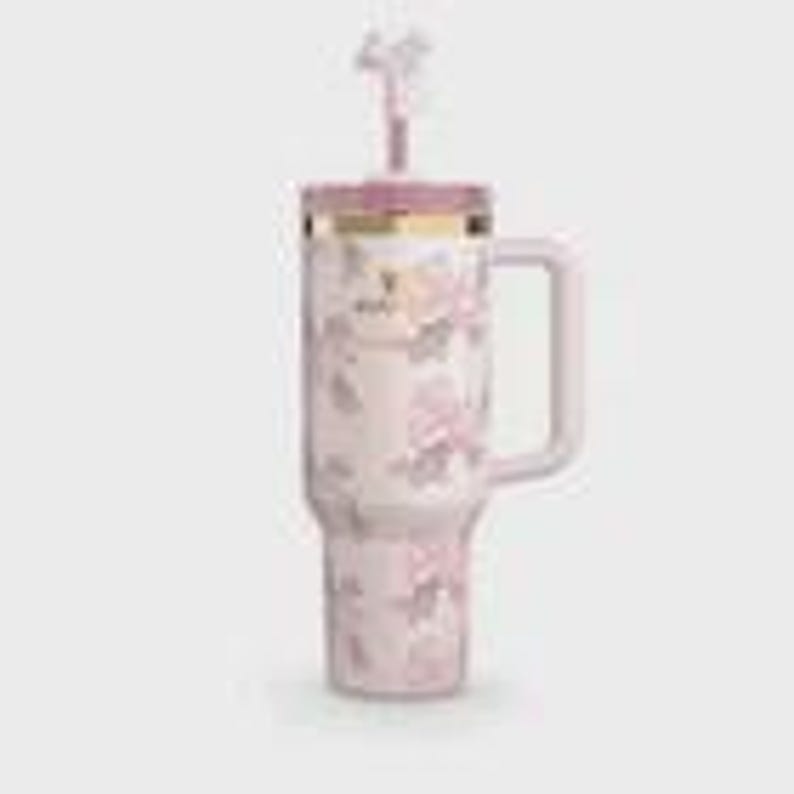 Puede incluir: Un vaso grande y aislado con asa, con un estampado floral en tonos rosa y blanco. Incluye una tapa y pajita a juego, con detalles dorados. El dise&ntilde;o del vaso presenta un elemento decorativo en la pajita.