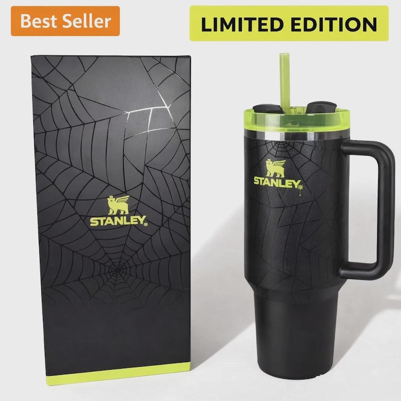 May include: A black Stanley travel mug with a green lid and straw, decorated with a spiderweb pattern. The mug has a handle and the Stanley logo. A matching black box with the same design is next to it. Text reads "Best Seller" and "Limited Edition."