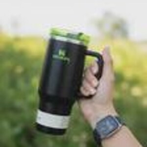 May include: Black insulated travel mug with a green lid and handle. The mug has a white band near the bottom and a green logo. The mug is held in a hand, with a watch visible on the wrist.
