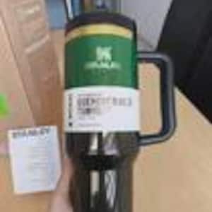 May include: A black Stanley Quencher travel mug with a large handle and a green and yellow band around the middle. The mug has a black lid and a white label with text. A cardboard box is in the background.