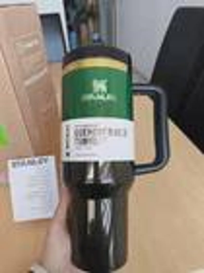 May include: A black Stanley Quencher travel mug with a large handle and a green and yellow band around the middle. The mug has a black lid and a white label with text. A cardboard box is in the background.