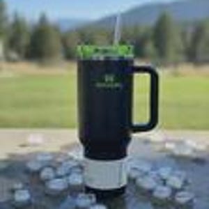 May include: Black and green insulated travel mug with a handle and a straw. The mug has a white base and a green lid. The mug has the text "Green Life" printed on the side. The mug is sitting on a surface with ice cubes scattered around it.