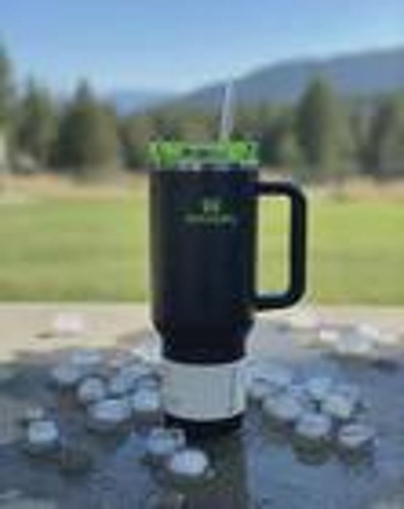 May include: Black and green insulated travel mug with a handle and a straw. The mug has a white base and a green lid. The mug has the text "Green Life" printed on the side. The mug is sitting on a surface with ice cubes scattered around it.