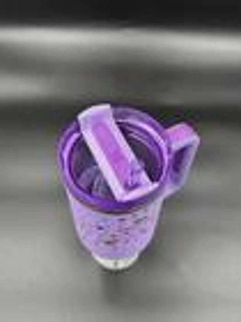 May include: Purple insulated travel mug with a handle and a flip-top lid. The mug has a speckled design and a white base. The lid is open, revealing the interior. The mug is designed to keep beverages hot or cold.