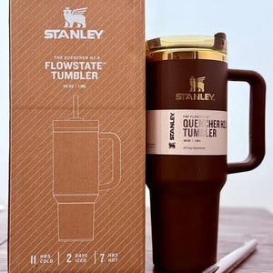 May include: A brown Stanley Flowstate tumbler with a handle and a gold-colored lid, next to its cardboard box. The box features the Stanley logo and product details, including "40 OZ | 1.18L". The tumbler is designed for all-day hydration.