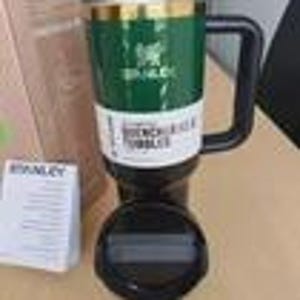 May include: A green and gold Stanley mug with a black handle and base. The mug has the Stanley logo and the words "French Press & Tumbler" on a white label. A white card is visible in the background.
