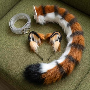 Puede incluir: Un conjunto de accesorios de piel sintética que incluye una cola larga a rayas con pelo naranja, blanco y negro, dos orejas a juego con mechones negros y naranjas, y un cinturón de plástico transparente con una hebilla de metal plateado.