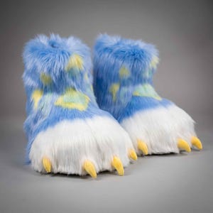 Puede incluir: Un par de pantuflas de felpa con pelo sintético largo y desgreñado en color azul brillante con manchas amarillas y verde claro. La parte delantera de cada pantufla está cubierta de pelo blanco e incluye tres garras de tela amarillas.