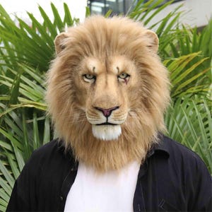 May include: A realistic lion mask with a tan mane and detailed facial features. The mask has a pink nose and green eyes. The person is wearing a white shirt and a black jacket. The background features green foliage.