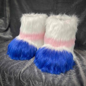 Puede incluir: Un par de botas hasta el tobillo cubiertas de piel sintética larga y peluda. Las botas presentan un diseño horizontal de bloques de color con una sección superior blanca, una banda central rosa claro y una base azul vibrante en el pie.