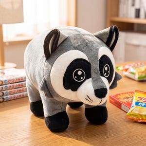 May include: A plush raccoon toy with soft grey fur, a white face mask, and black patches around the eyes. The stuffed animal features black ears, black paws, and embroidered eyes with white highlights, standing in a quadrupedal position.