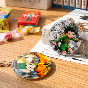 May include: Two circular acrylic keychains featuring anime-style character illustrations. One keychain displays a character in a green outfit against a black and white manga panel background. The other shows two characters with orange and green hair.