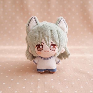 May include: A plush doll featuring soft sage green fur, pointed animal ears, and large brown embroidered eyes. The character has short, layered hair and wears a white short-sleeved shirt with blue trim and matching blue shorts, crafted with a smooth, rounded body shape.