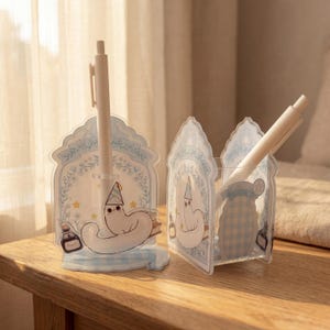 Puede incluir: Dos portalápices de acrílico transparente con una criatura blanca con gorro de fiesta, rodeada de ilustraciones florales azules y pequeñas estrellas amarillas. Cada soporte tiene un patrón de cuadros azul claro y un gráfico de un tintero.